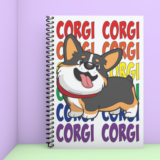 A journal leaning in a corner. the journal has a tricolor corgi on the front. The text behind the corgi says "CORGI CORGI". The top lines of text are red and each row is another color of the rainbow. 