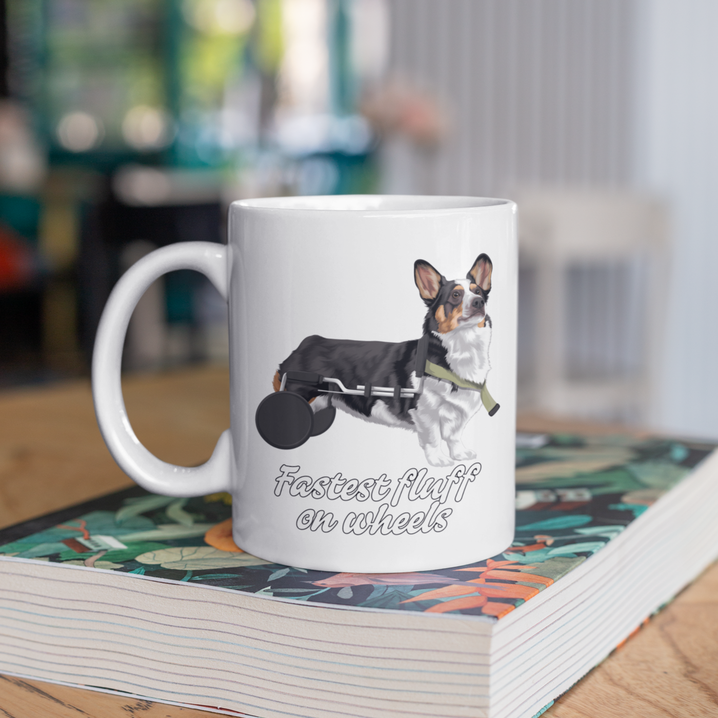 A white coffee mug sitting on a book on a table. The design on the mug is a tricolor corgi that is using a dog wheelchair. There is black-outlined white text that says "Fastest fluff on wheels". 