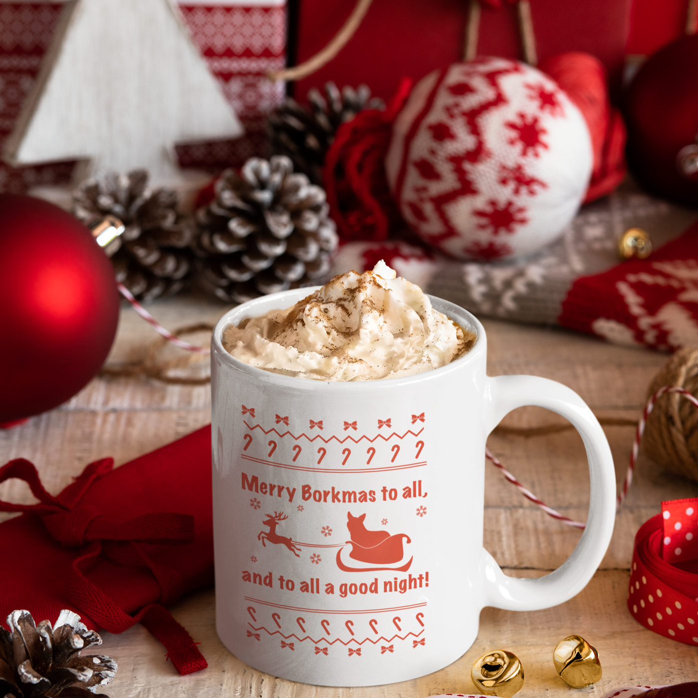 A white mug with the text "Merry Borkmas to all, and to all a good night!" with a Christmas pattern, and a corgi in Santa's sleigh. The white mug is sitting amongst a number of Christmas decorations. 