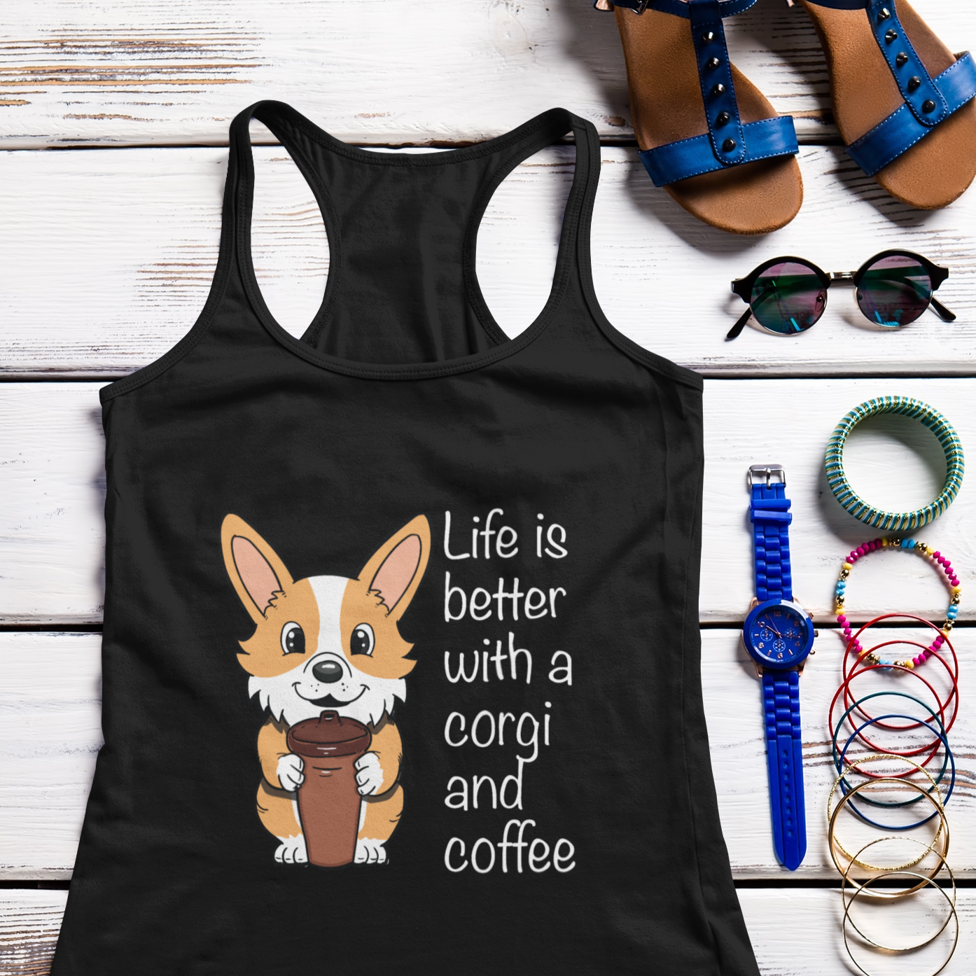 A flat lay of a racerback tank top that is black. The design of the tank is a fawn corgi holding a travel coffee mug. The text next to the design says "Life is better with a corgi and coffee." There's some shoes and accessories next to the tank top. 