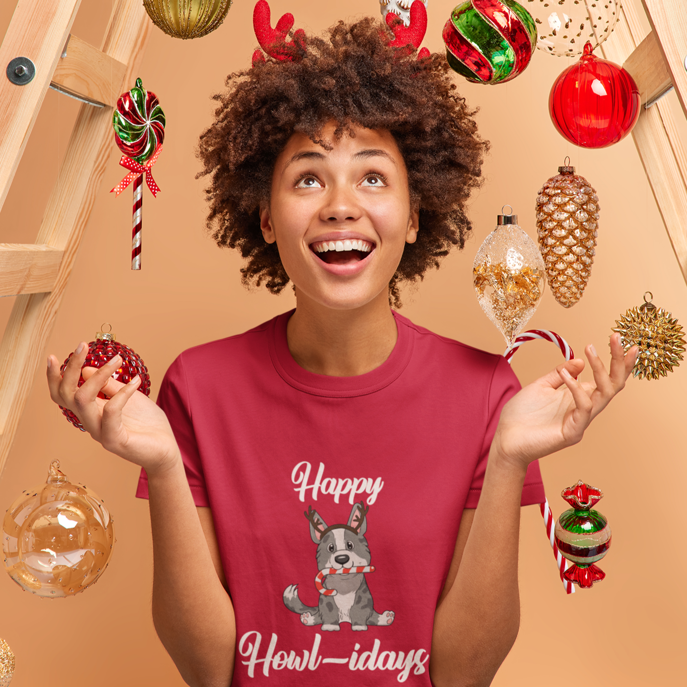 A model wearing a red shirt that has a cardigan corgi on it. The corgi is wearing fake reindeer horns and is holding a candy cane in its mouth. The text says "Happy Howl-idays".