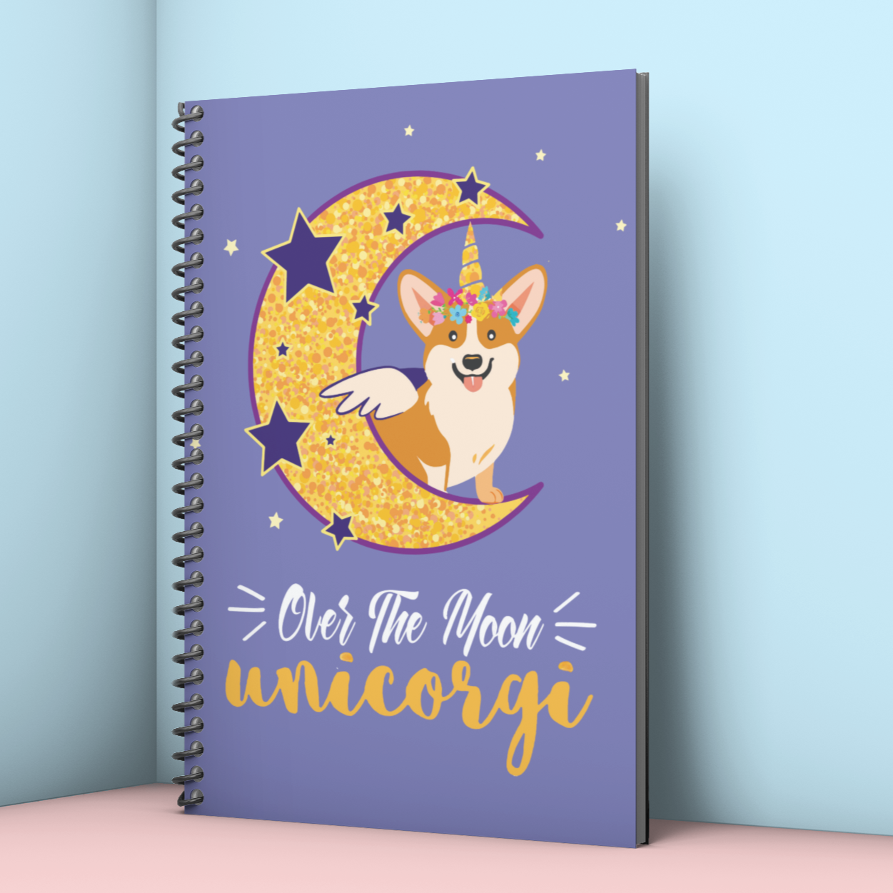 A spiral bound notebook leaning in a corner with a blue background, a corgi with a unicorn horn and wings sitting on the moon. The text says "Over the moon Unicorgi". 