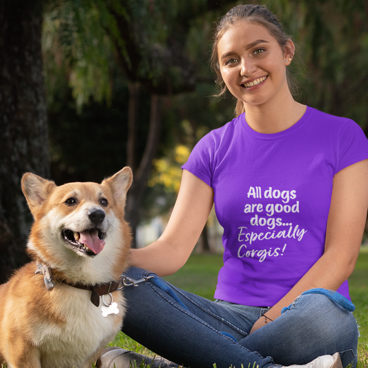 A model wearing a purple short sleeved shirt. The white text on the shirt says "All dogs are good dogs... Especially Corgis!". Sitting next to the model is a Welsh Pembroke Corgi. 