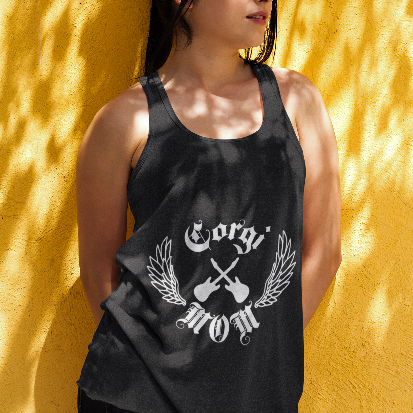 A woman leaning up against the wall in dappled sunlight. She's wearing a black racerbank tank top that says "Corgi Mom" in old english type with crossed guitars and wings. 